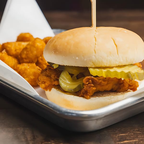 A fried chicken sandwich with pickles and tater tots on a metal tray.