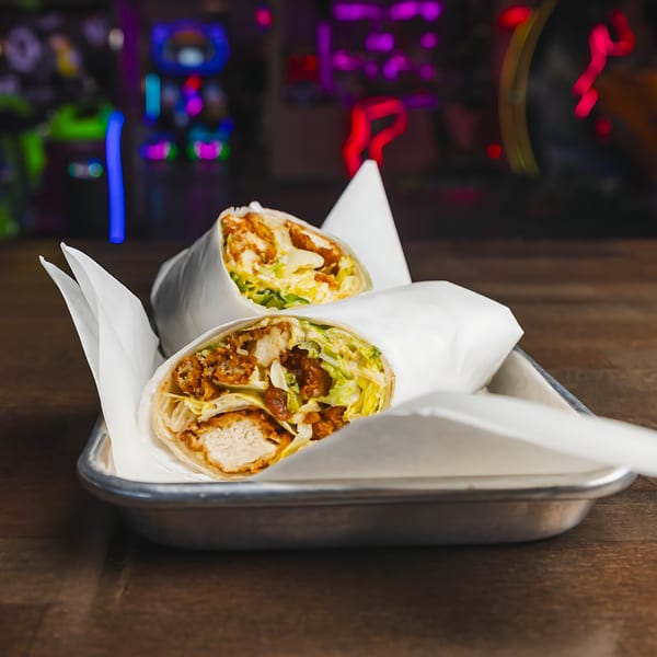 Two IWingz chicken wraps with lettuce in a metal tray, against a blurred background of neon lights.