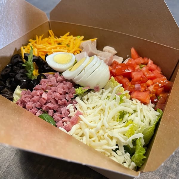 A colorful chef salad with ham, turkey, cheese, olives, tomatoes, and hard-boiled eggs in a to-go box.
