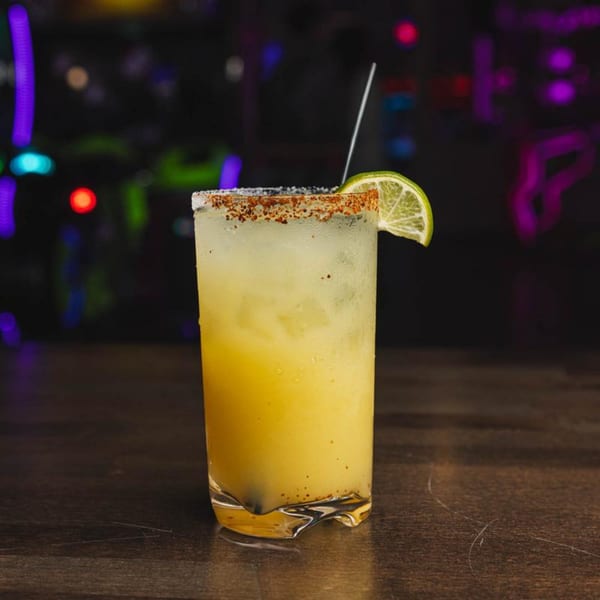 A tall glass of yellow IWingz drink with a salted rim and lime wedge on a dark table.