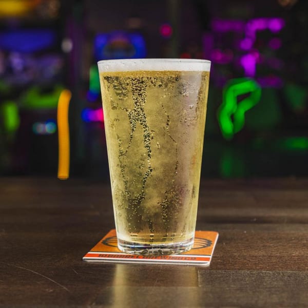 A tall glass of bubbly, golden beer on a coaster, with neon lights blurred in the background.